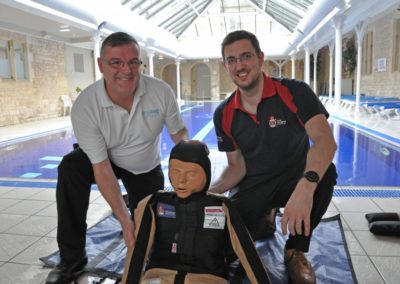 Two men with a training dummy by a pool.