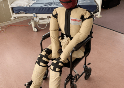 Medical training dummy in a wheelchair next to hospital bed.