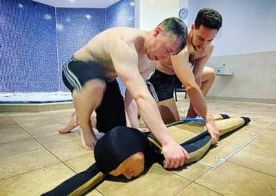Men practising rescue techniques with dummy in indoor pool area.